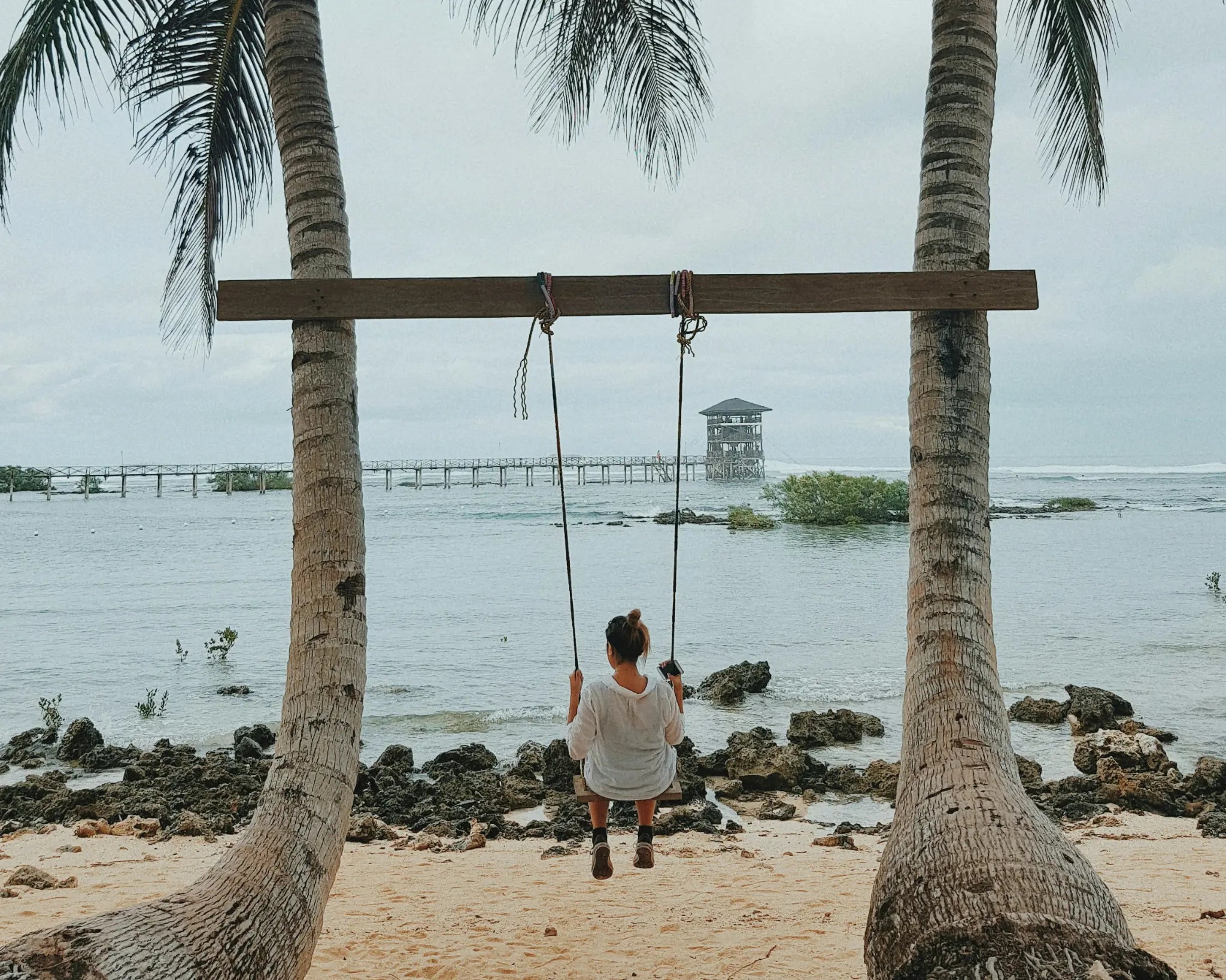 A traveller on a swing between palm trees facing the sea